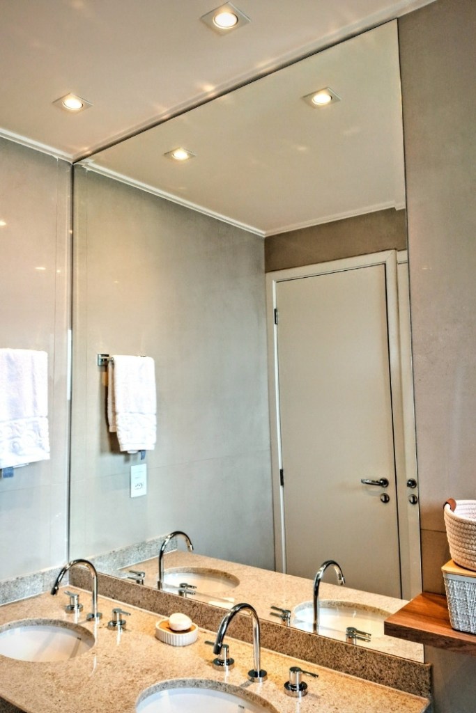 Espelho grande ocupando parede inteira acima de bancada de granito com duas cubas no banheiro.